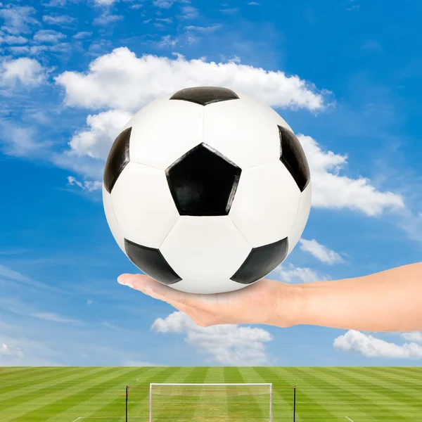 Pelota de fútbol en la mano con campo de fútbol y cielo azul — Foto de Stock