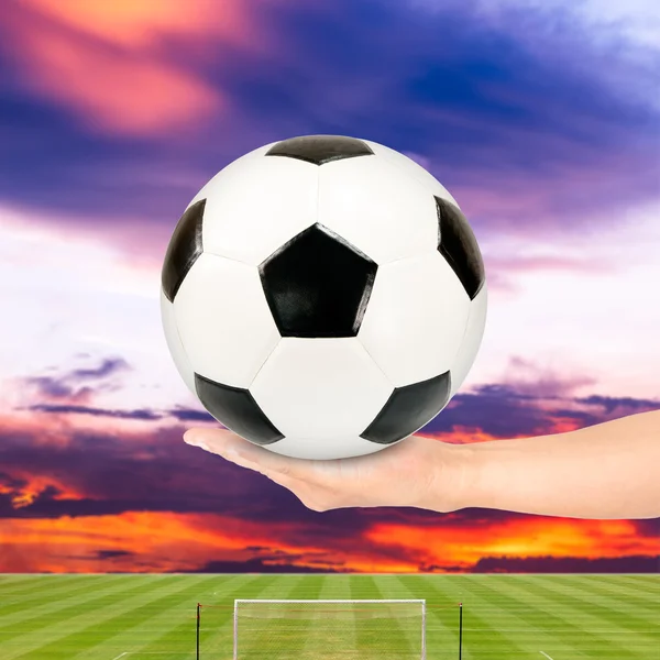 Soccer ball in hand with soccer field and sunset sky background — Stock Photo, Image
