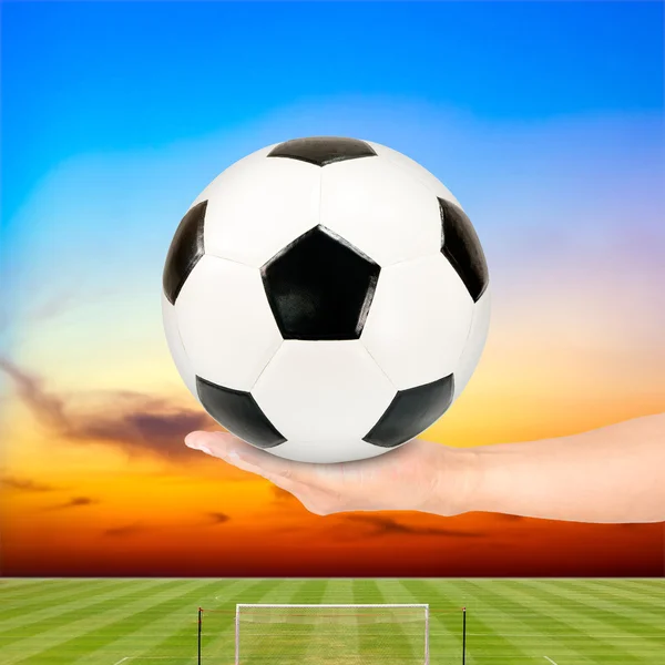 Fußball in der Hand mit Fußballfeld und Sonnenuntergang Himmel Hintergrund — Stockfoto