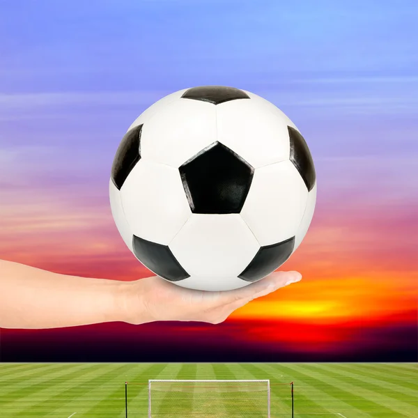 Fußball in der Hand mit Fußballfeld und Sonnenuntergang Himmel Hintergrund — Stockfoto