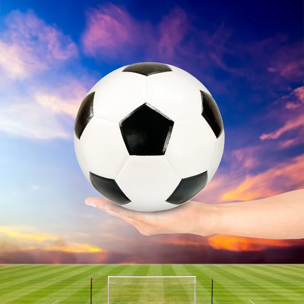 Fußball in der Hand mit Fußballfeld und Sonnenuntergang Himmel Hintergrund — Stockfoto