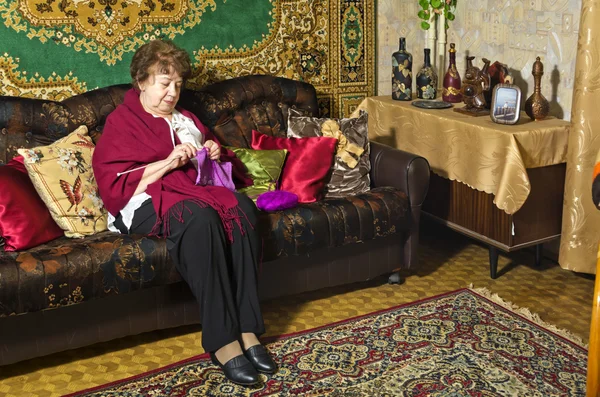 Una donna anziana seduta su un divano e lavora a maglia — Foto Stock