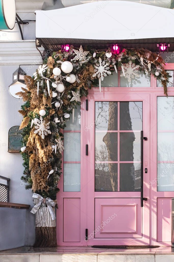 Winter festive decoration of a pink door for the New Year with branches of a Christmas tree, snowflakes, toys, bells and garlands. Christmas composition for home decoration for winter holidays