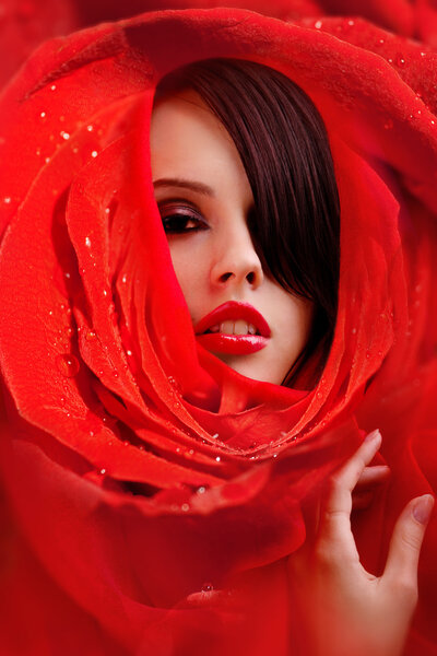 Wrapped in red roses petals beautiful young female face with a glamorous make-up with glossy red lips