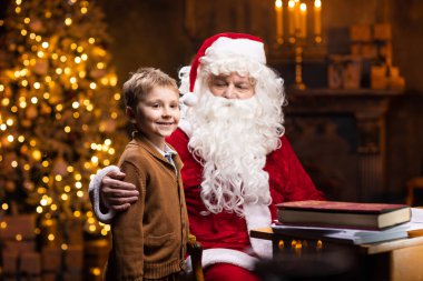 Noel Baba ve küçük çocuk. Neşeli Noel Baba masada otururken çalışıyor. Arka planda şömine ve Noel ağacı var. Noel konsepti.