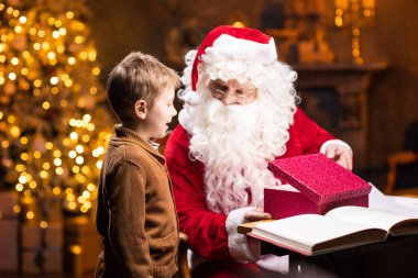 Noel Baba ve küçük çocuk. Neşeli Noel Baba masada otururken çalışıyor. Arka planda şömine ve Noel ağacı var. Noel konsepti.