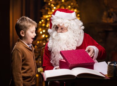 Noel Baba ve küçük çocuk. Neşeli Noel Baba masada otururken çalışıyor. Arka planda şömine ve Noel ağacı var. Noel konsepti.