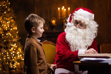Noel Baba ve küçük çocuk. Neşeli Noel Baba masada otururken çalışıyor. Arka planda şömine ve Noel ağacı var. Noel konsepti.