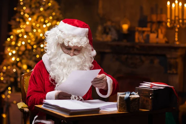Lieu de travail du Père Noël. Le Père Noël joyeux lit les lettres des enfants assis à la table. Cheminée et sapin de Noël en arrière-plan. Concept de Noël. — Photo