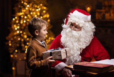 Noel Baba ve küçük çocuk. Neşeli Noel Baba masada otururken çalışıyor. Arka planda şömine ve Noel ağacı var. Noel konsepti.