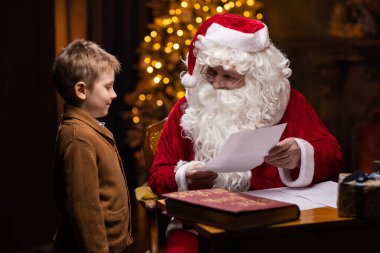 Noel Baba ve küçük çocuk. Neşeli Noel Baba masada otururken çalışıyor. Arka planda şömine ve Noel ağacı var. Noel konsepti.