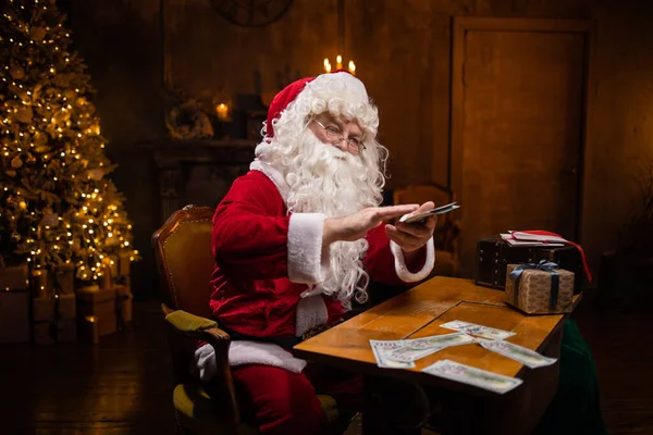サンタクロースの職場。陽気なサンタはテーブルに座ってお金を数えている。背景にある暖炉とクリスマスツリー。クリスマスのコンセプト. — ストック写真
