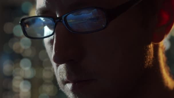 コンピュータで働いている若い男のクローズアップ肖像画。ノートパソコンのディスプレイはメガネに反映されます。背景にナイトシティライト。ビジネス、コーディング、交換の概念. — ストック動画