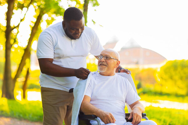 Caregiver and old man in a wheelchair. Professional nurse and patient walking outdoor in the park at sunset. Assistance, rehabilitation and health care.