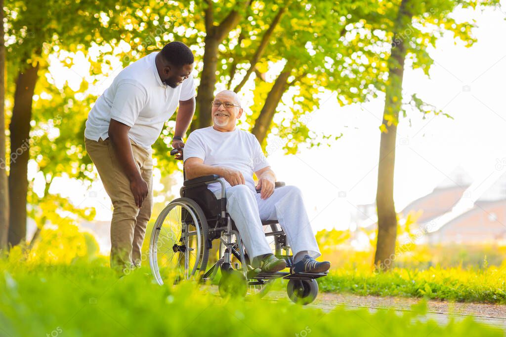 Caregiver and old man in a wheelchair. Professional nurse and patient walking outdoor in the park at sunset. Assistance, rehabilitation and health care.