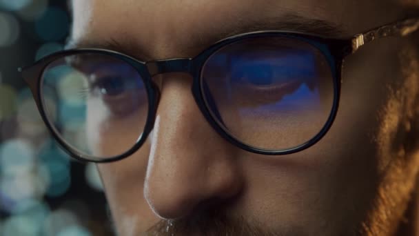 Retrato de cerca de un joven que trabaja en la computadora. La pantalla del ordenador portátil se refleja en las gafas. Luces nocturnas en el fondo. Concepto de negocio, codificación e intercambio. — Vídeos de Stock