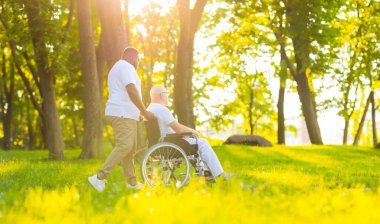 Bakıcı ve tekerlekli sandalyedeki yaşlı adam. Profesyonel hemşire ve hasta gün batımında parkta yürüyorlar. Yardım, rehabilitasyon ve sağlık hizmetleri.