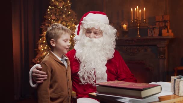 Święty Mikołaj i mały chłopiec. Wesoły Mikołaj pracuje siedząc przy stole. Kominek i choinka w tle. Koncepcja świąteczna. — Wideo stockowe