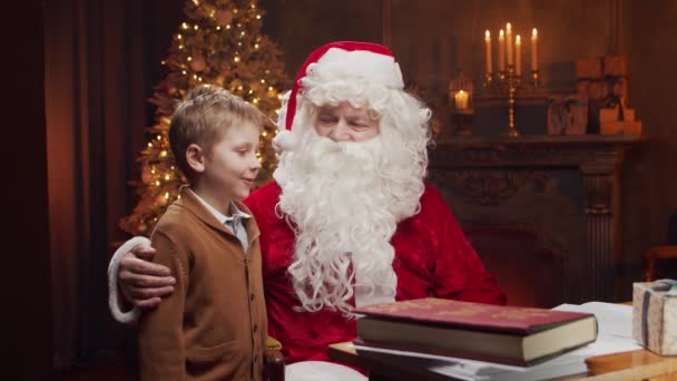 Père Noël et petit garçon. Père Noël joyeux travaille tout en étant assis à la table. Cheminée et sapin de Noël en arrière-plan. Concept de Noël. — Video