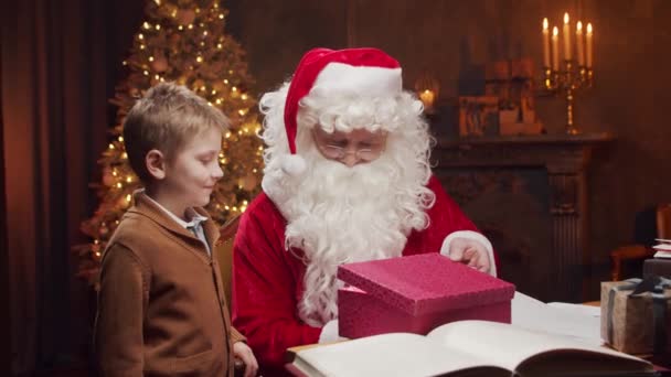 Père Noël et petit garçon. Père Noël joyeux travaille tout en étant assis à la table. Cheminée et sapin de Noël en arrière-plan. Concept de Noël. — Video