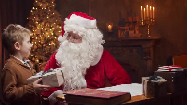 Santa Claus y el niño pequeño. Alegre Santa está trabajando mientras está sentado a la mesa. Chimenea y árbol de Navidad en el fondo. Concepto de Navidad. — Vídeo de stock