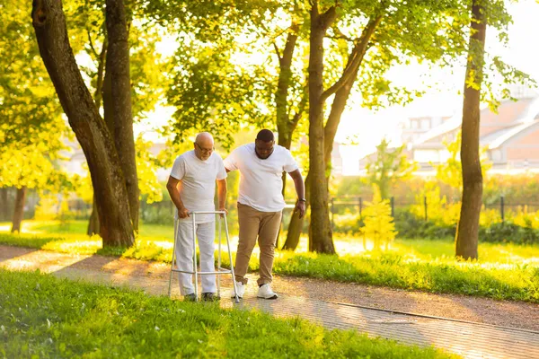 Caregiver mengajarkan orang tua berjalan dengan walker. Perawat profesional dan pasien berjalan keluar rumah saat matahari terbenam. Bantuan, rehabilitasi dan perawatan kesehatan. — Stok Foto