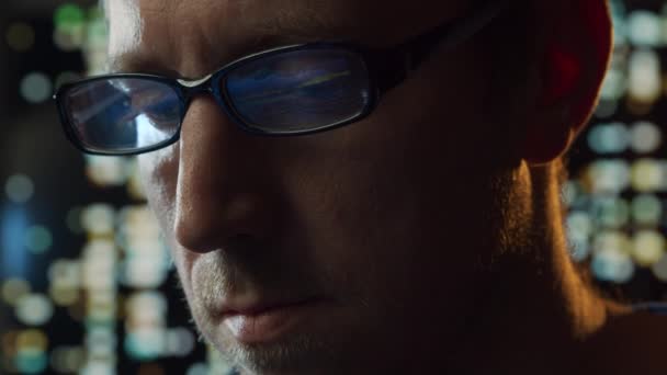 Közelkép egy fiatalemberről, aki a számítógépnél dolgozik. A laptop kijelzője tükröződik a szemüvegben. Éjszakai fények a háttérben. Üzleti, kódolási és cserekoncepció. — Stock videók
