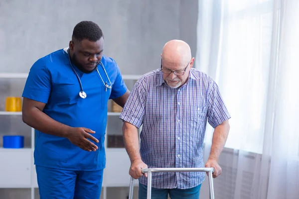 Caregiver is teaching old man to walk with walker. Professional nurse and patient in a nursing home. Assistance, rehabilitation and health.