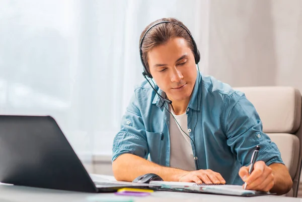 Un joven con auriculares trabaja en una computadora portátil. Freelancer, trabajador remoto o lugar de trabajo estudiantil. Concepto de trabajo distante. —  Fotos de Stock