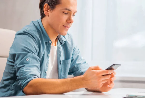 Un joven con un smartphone se sienta a la mesa en casa. Freelancer, trabajador remoto o lugar de trabajo estudiantil. Concepto de trabajo distante. — Foto de Stock