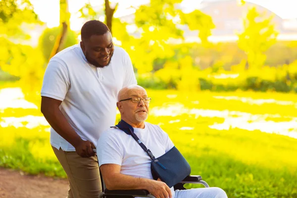 Cuidador y anciano en silla de ruedas. Enfermera profesional y paciente caminando al aire libre en el parque al atardecer. Asistencia, rehabilitación y asistencia sanitaria. —  Fotos de Stock