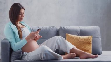 Genç hamile kadın elinde cep telefonuyla evde dinleniyor ve bebek bekliyor. Hamilelik, annelik, sağlık ve yaşam tarzı kavramı.