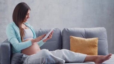Genç hamile bir kadın tablet bilgisayarla evde dinleniyor ve bebek bekliyor. Hamilelik, annelik, sağlık ve yaşam tarzı kavramı.