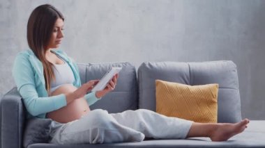 Genç hamile bir kadın tablet bilgisayarla evde dinleniyor ve bebek bekliyor. Hamilelik, annelik, sağlık ve yaşam tarzı kavramı.
