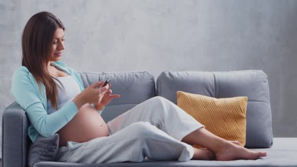 妊娠中の若い女性はスマートフォンで自宅で休んでおり、赤ちゃんを期待しています。妊娠、母親、健康、ライフスタイルの概念. — ストック動画