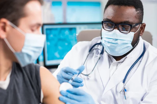 Afroamerikansk läkare tillverkar ett coronavirusvaccin med hjälp av en spruta och injektionsnål. Professionell läkare och patient på sjukhuskontoret. Vaccination och säkerhet. — Stockfoto