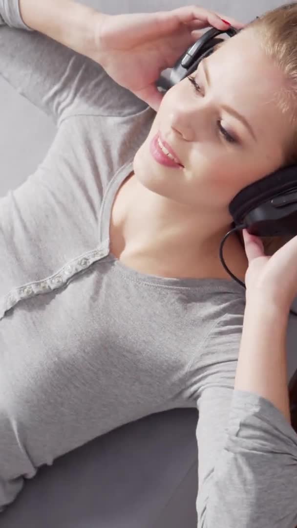 Ung vacker flicka ligger på en soffa i hörlurar och lyssnar på musik. Blond kvinna vilar hemma. — Stockvideo