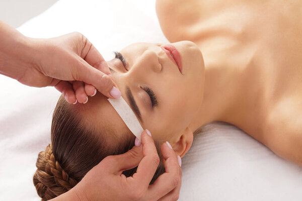 Beautician is removing hair from beautiful female face with hot wax. Woman has a beauty treament procedure. Depilation, epilation, skin and health care concepts.