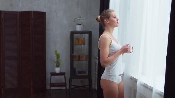 Ung flicka står framför fönstret och tittar in i det. Blond kvinna hemma i vardagsrummet. — Stockvideo