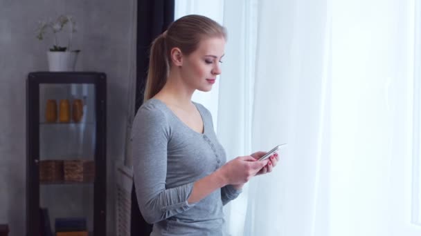 若い十代の女の子は窓の前にスマートフォンで立っているとそれを調べています。自宅のリビングで女性. — ストック動画