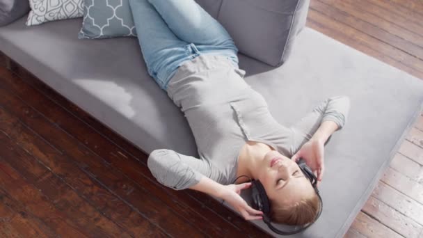 Joven chica hermosa se encuentra en un sofá en los auriculares y escucha música. Mujer rubia está descansando en casa. — Vídeos de Stock