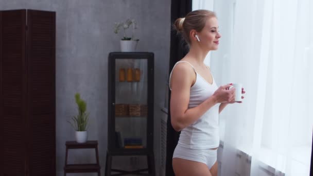 Mujer joven en ropa interior blanca está de pie delante de la ventana y mirando dentro de ella. Chica en casa en la sala de estar. — Vídeos de Stock