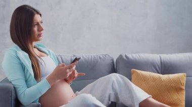 Genç hamile kadın elinde cep telefonuyla evde dinleniyor ve bebek bekliyor. Hamilelik, annelik, sağlık ve yaşam tarzı kavramı.