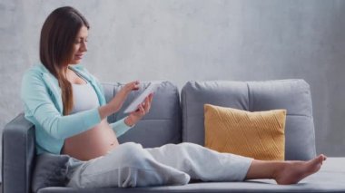 Genç hamile bir kadın tablet bilgisayarla evde dinleniyor ve bebek bekliyor. Hamilelik, annelik, sağlık ve yaşam tarzı kavramı.