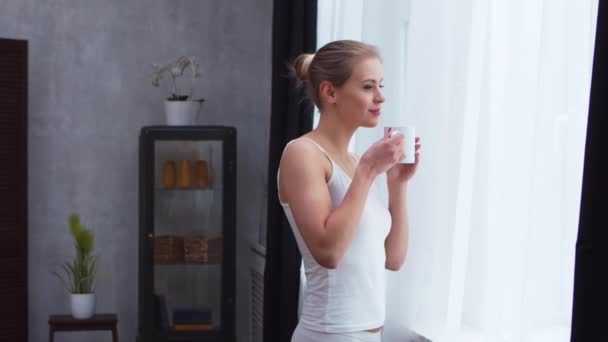 Ung kvinna i vita underkläder står framför fönstret och tittar in i det. Flicka hemma i vardagsrummet. — Stockvideo