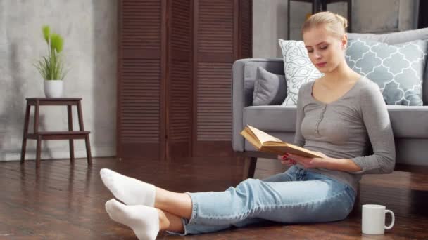 Jovem loira está lendo um livro ou um livro didático. Uma mulher bonita está descansando e relaxando em casa na sala de estar. — Vídeo de Stock