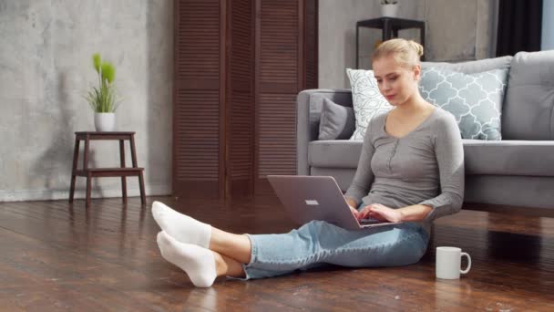 Egy fiatal nő laptoppal dolgozik, miközben otthon ül a padlón. Diák, vállalkozó vagy szabadúszó lány, aki távolról dolgozik vagy tanul. Távolsági tanulás és munkahely fogalma. — Stock videók