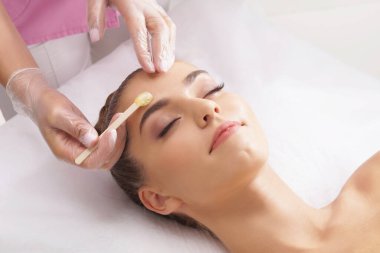 Güzellik uzmanı, güzel kadın yüzündeki kılları sıcak balmumuyla alıyor. Kadının güzel bir titreme prosedürü var. Defilasyon, epilasyon, cilt ve sağlık konseptleri.
