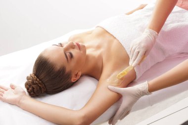 Güzellik uzmanı, genç kadın koltukaltlarındaki kılları sıcak balmumuyla alıyor. Kadının güzel bir titreme prosedürü var. Defilasyon, epilasyon, cilt ve sağlık konseptleri.