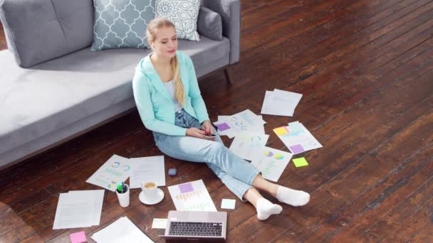 Mujer joven trabaja con documentos utilizando un ordenador portátil mientras está sentado en el suelo en casa. Estudiante, empresaria o freelancer que trabaja o estudia a distancia. Concepto de aprendizaje a distancia y empleo. — Vídeos de Stock